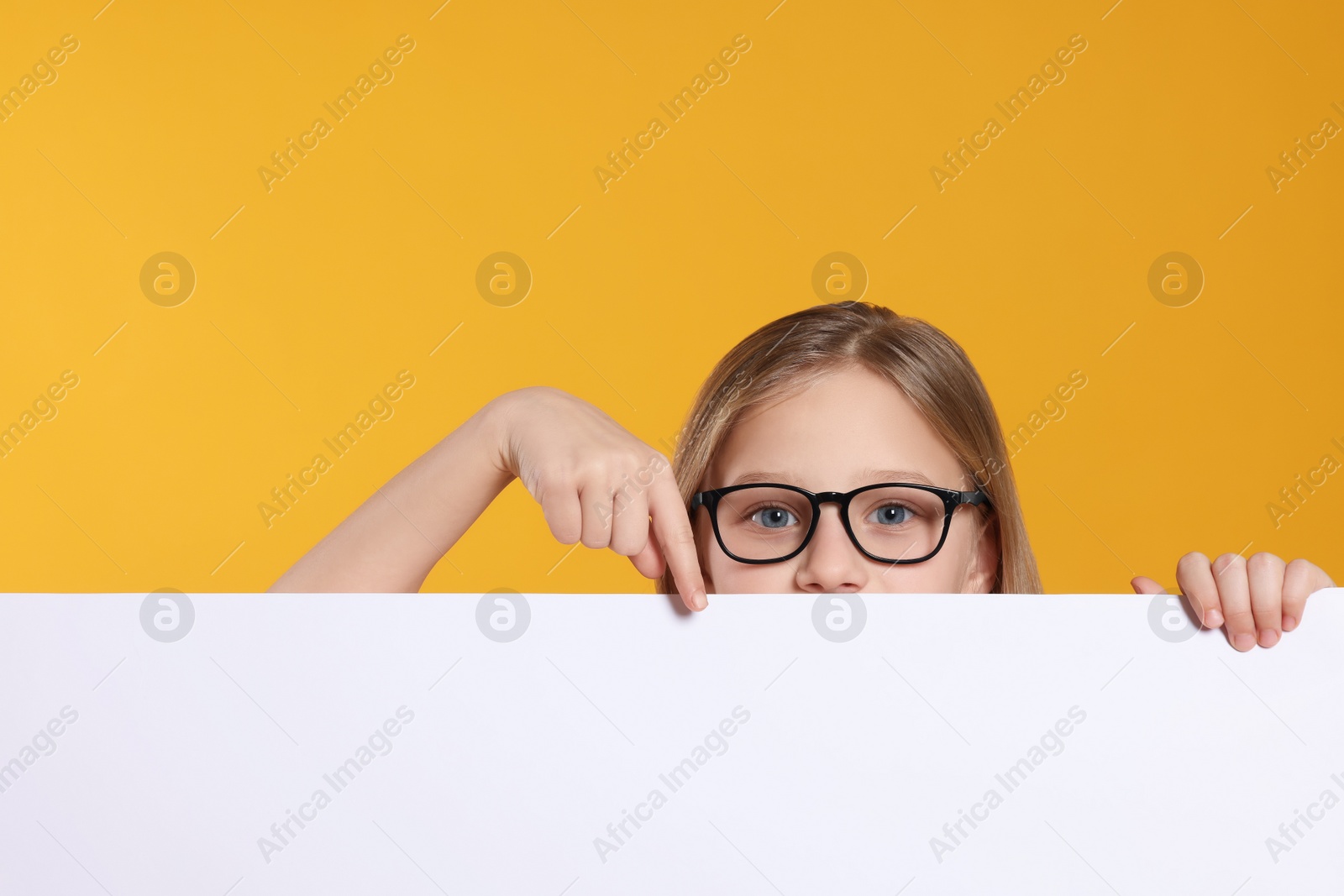 Photo of Cute girl looking out of placard and pointing on it against orange background. Space for text