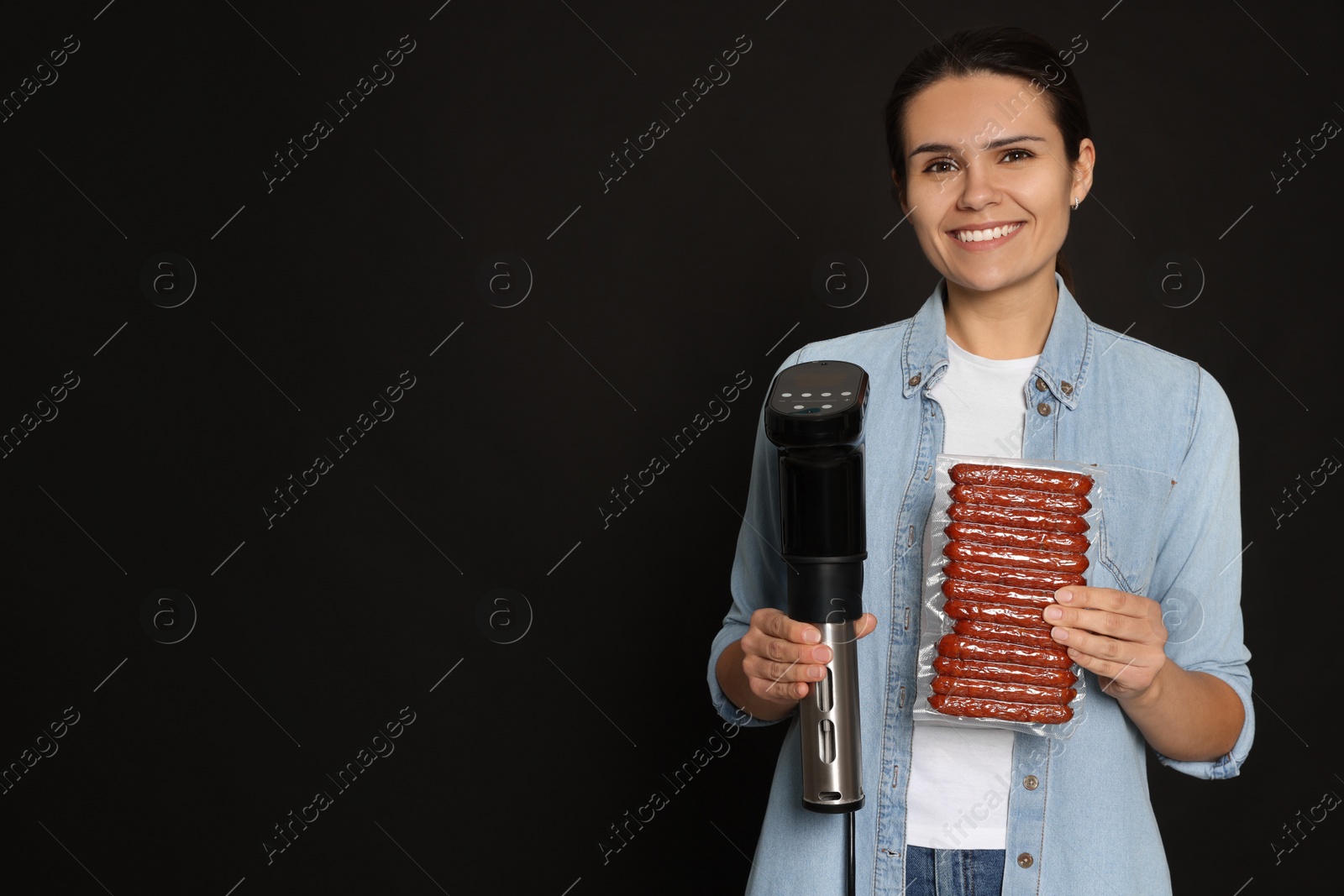 Photo of Beautiful young woman holding sous vide cooker and sausages in vacuum pack on black background. Space for text