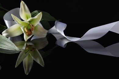 Photo of White lily and ribbon on black mirror surface in darkness, closeup with space for text. Funeral symbols