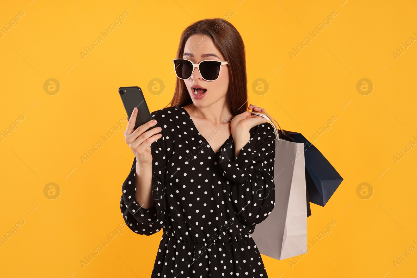 Photo of Surprised young woman with shopping bags looking at smartphone on yellow background. Big sale