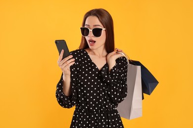 Photo of Surprised young woman with shopping bags looking at smartphone on yellow background. Big sale