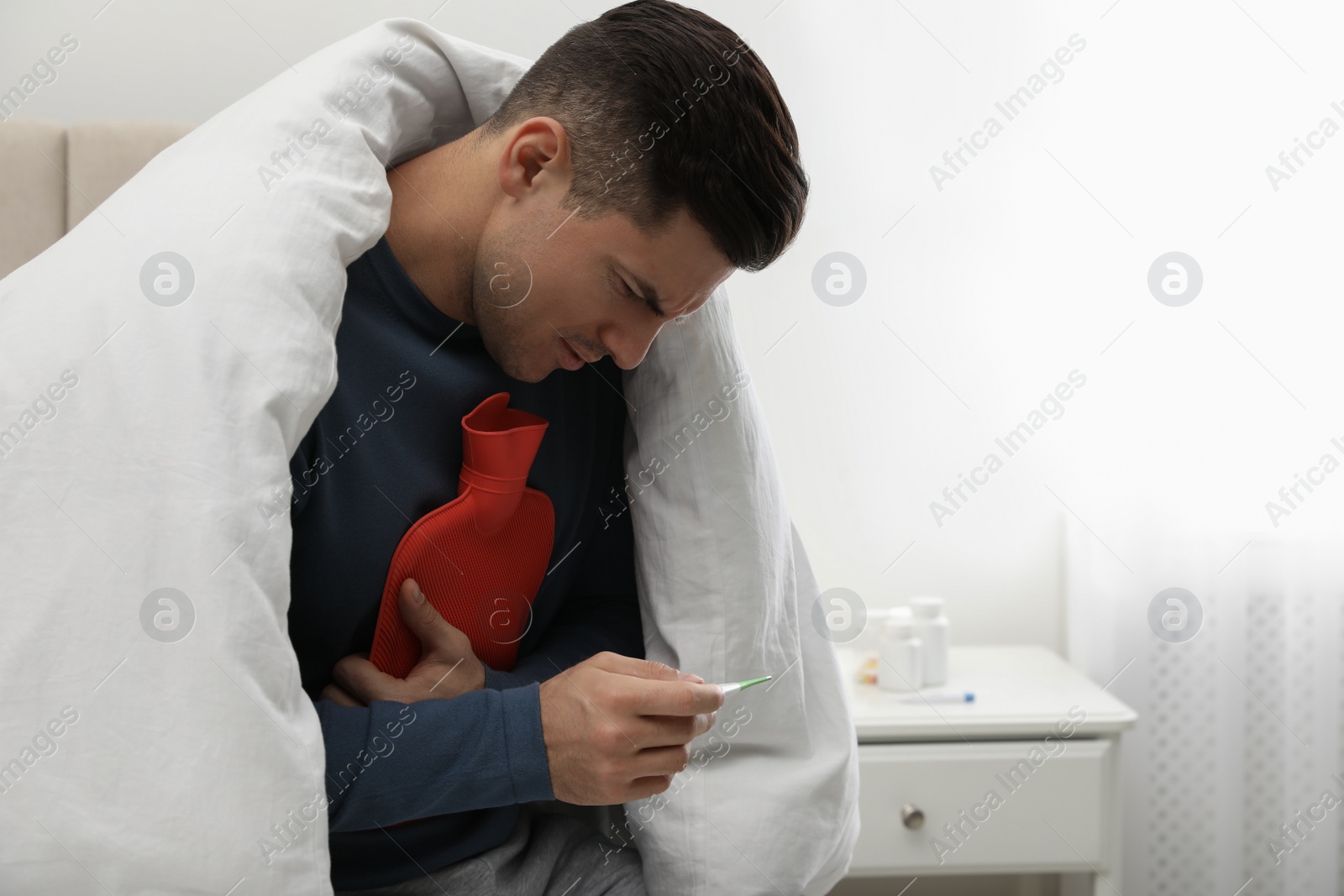 Photo of Ill man with hot water bottle checking his temperature at home