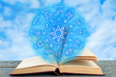 Old book on wooden table and illustration of zodiac wheel with astrological signs against blue sky