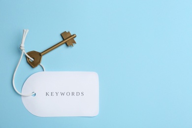 Photo of Metal key and tag wIth word KEYWORDS on light blue background, top view. Space for text
