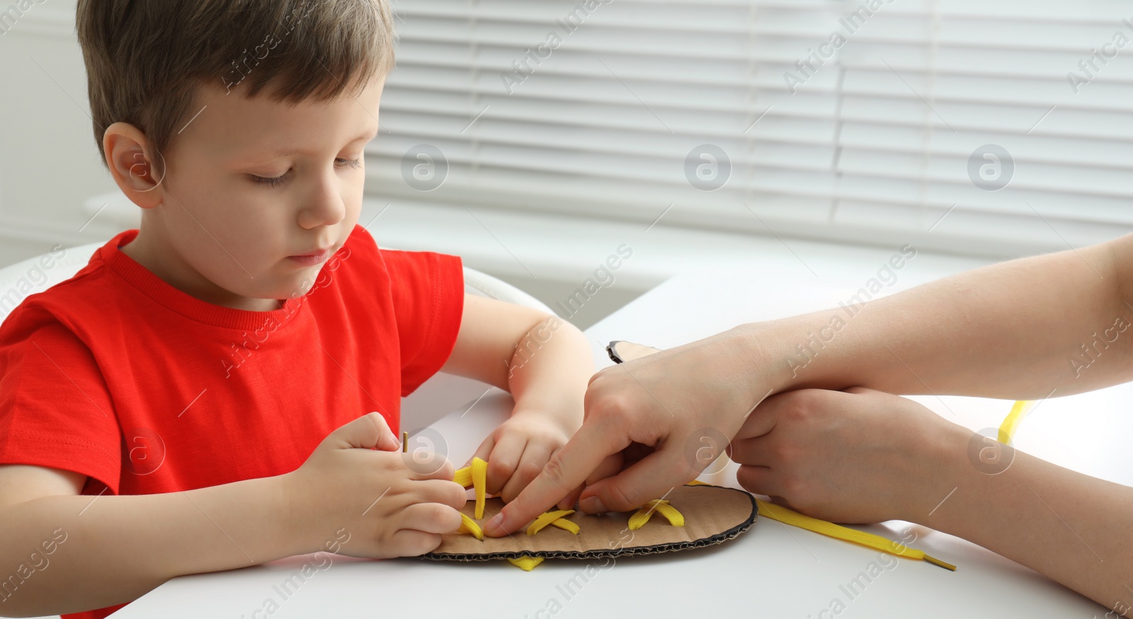 Photo of Mother teaching son to tie shoe laces using training cardboard template at white table, closeup