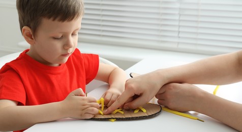 Mother teaching son to tie shoe laces using training cardboard template at white table, closeup