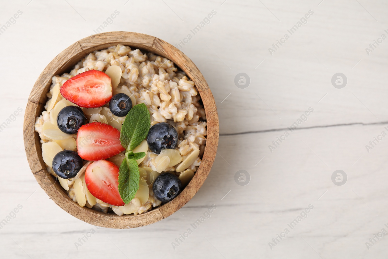 Photo of Tasty oatmeal with strawberries, blueberries and almond petals in bowl on white wooden table, top view. Space for text