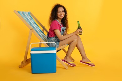 Photo of Happy young African American woman with bottle of beer resting in deck chair near cool box on yellow background