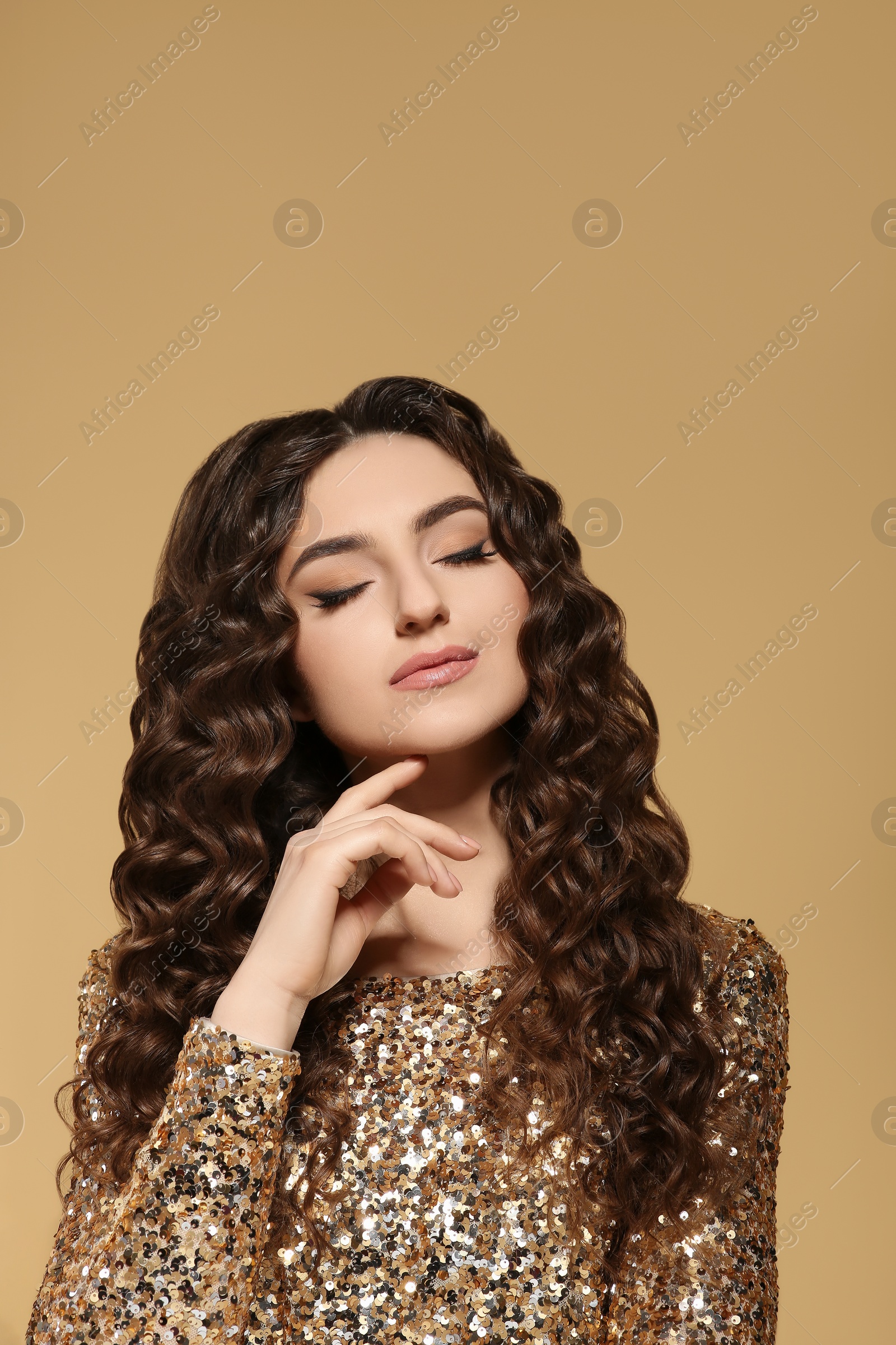 Photo of Beautiful young woman with long curly brown hair in golden sequin dress on beige background