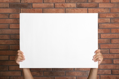 Man holding white blank poster near red brick wall, closeup. Mockup for design