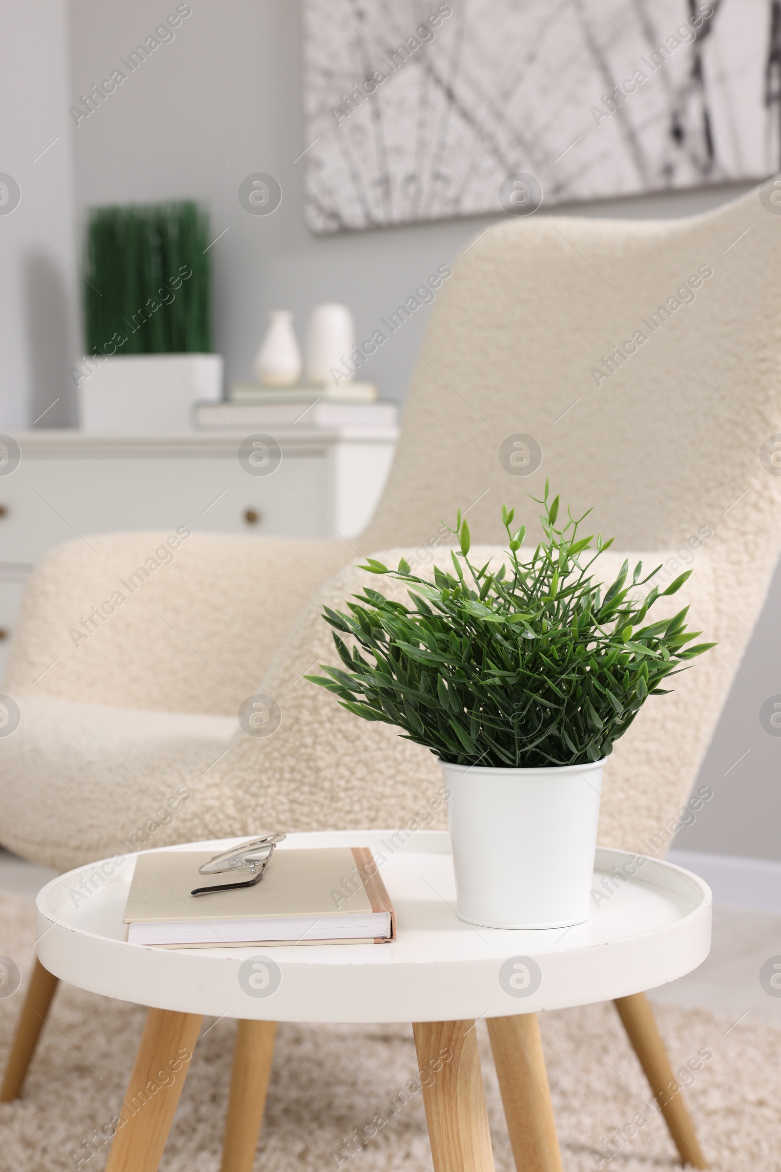 Photo of Potted artificial plant, book and glasses on side table near armchair indoors