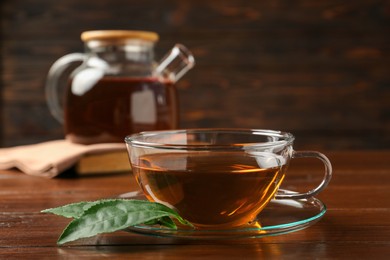 Photo of Cup of aromatic tea, green leaves and teapot on wooden table, closeup