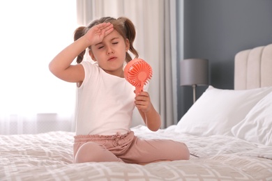 Little girl enjoying air flow from portable fan on bed in room. Summer heat