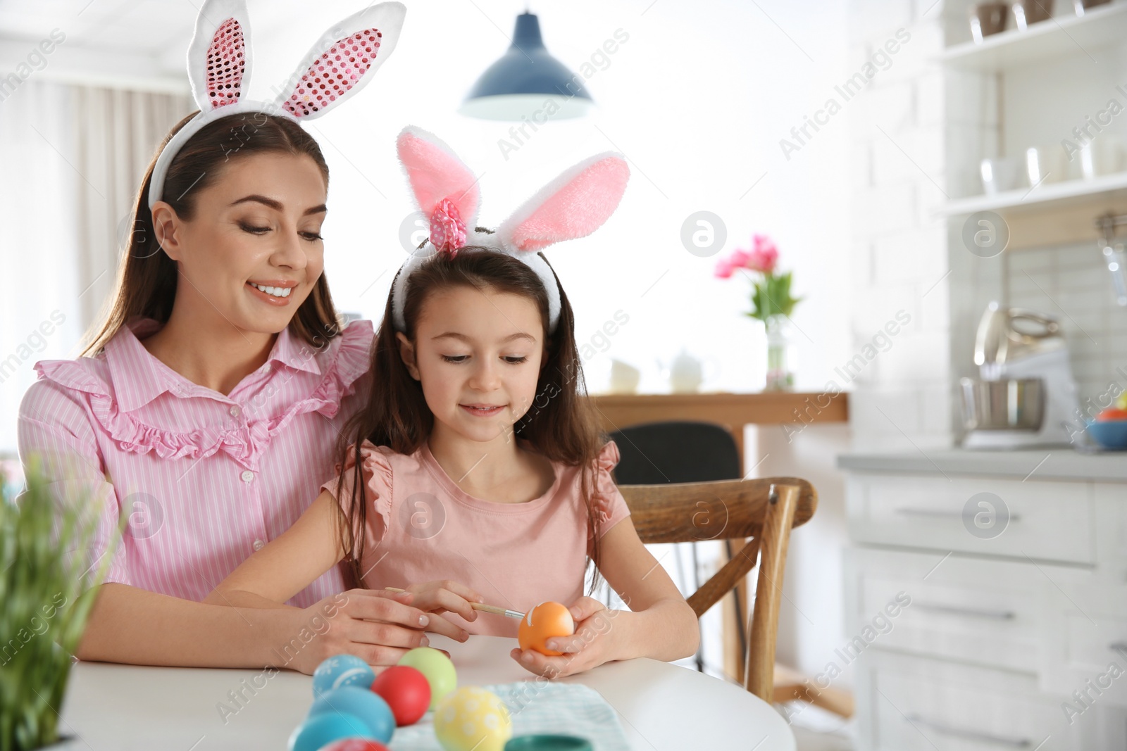 Photo of Mother and daughter with bunny ears headbands painting Easter eggs in kitchen, space for text