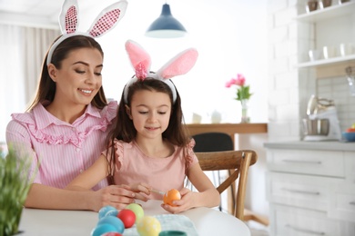 Mother and daughter with bunny ears headbands painting Easter eggs in kitchen, space for text