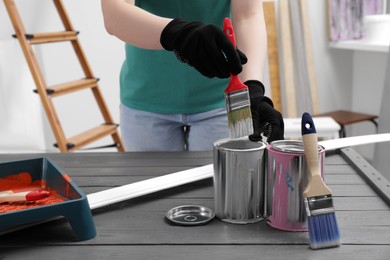 Photo of Woman dipping brush into can of white paint at black wooden table indoors, closeup