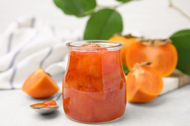 Jar of tasty persimmon jam and ingredients on white table, closeup