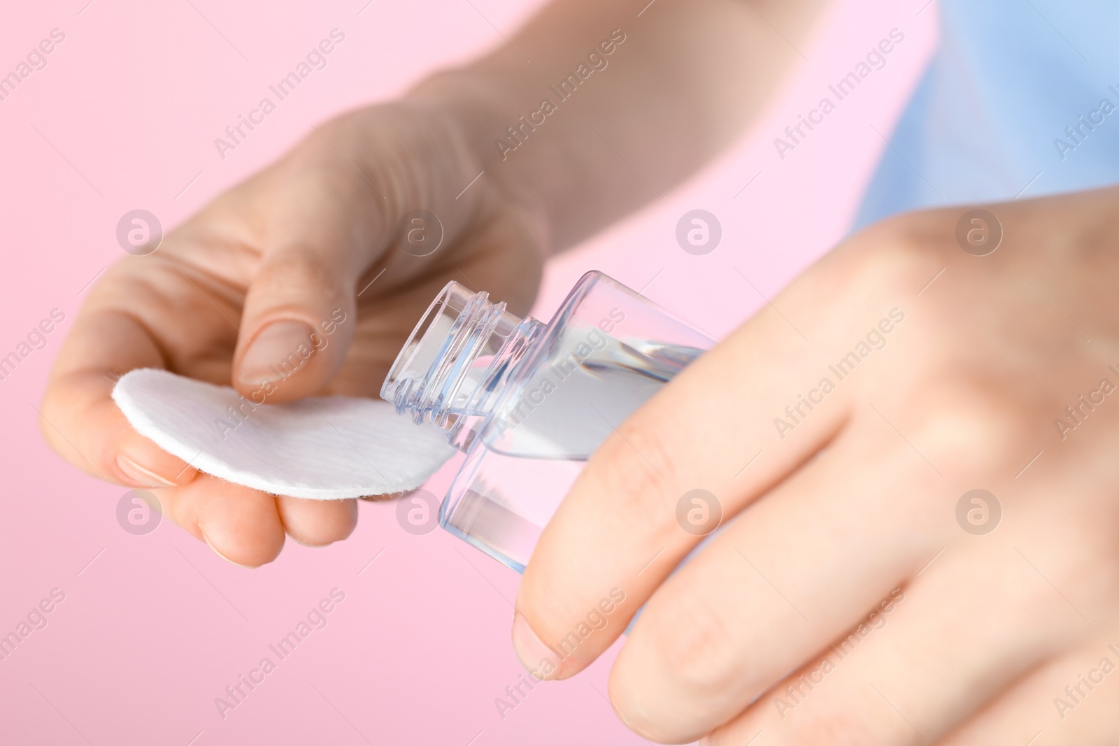 Photo of Woman pouring micellar water from bottle on cotton pad against pink background, closeup