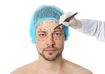 Doctor drawing marks on man's face for cosmetic surgery operation against white background