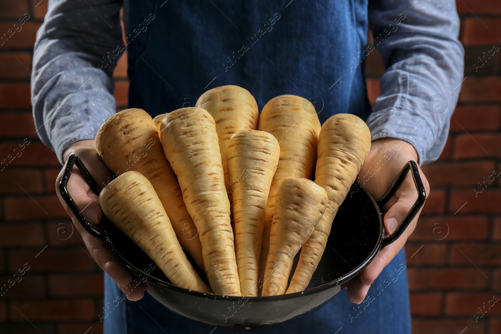 Photo of Woman holding bowl with fresh ripe parsnips near red brick wall, closeup