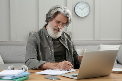 Photo of Middle aged man with laptop, notebook and cup of drink learning at table indoors