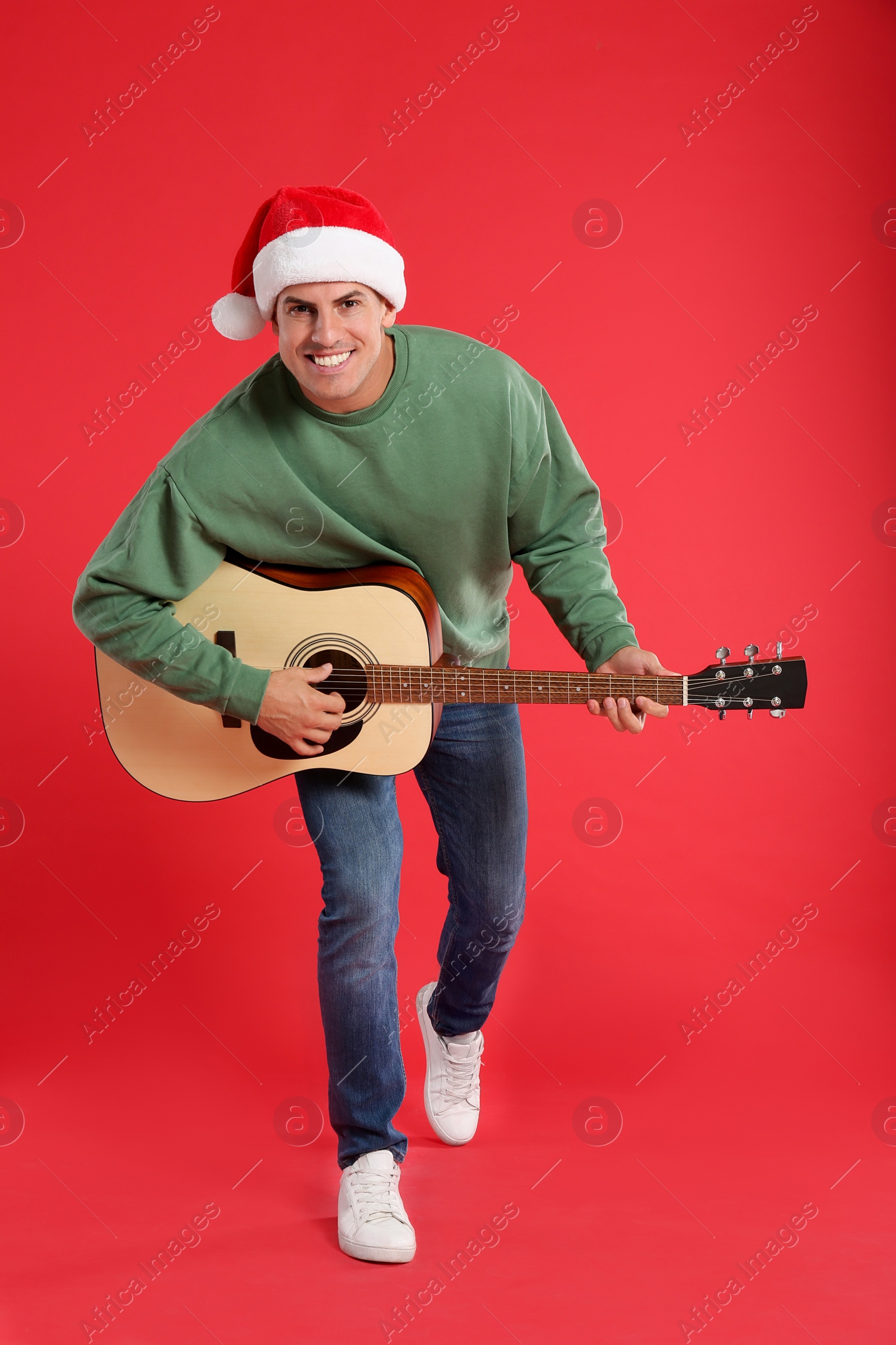 Photo of Man in Santa hat playing acoustic guitar on red background. Christmas music