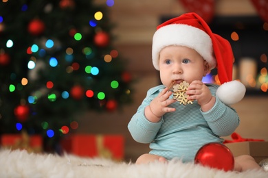 Photo of Little baby in Santa hat playing with Christmas decoration against blurred festive lights indoors