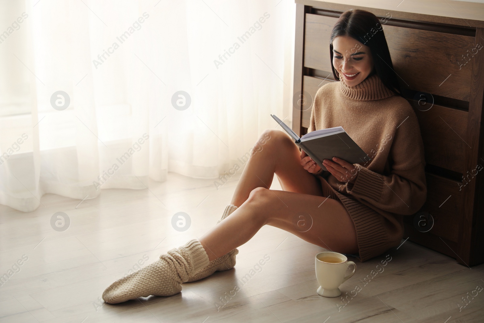 Photo of Woman with cup of drink and book sitting on warm floor at home. Heating system