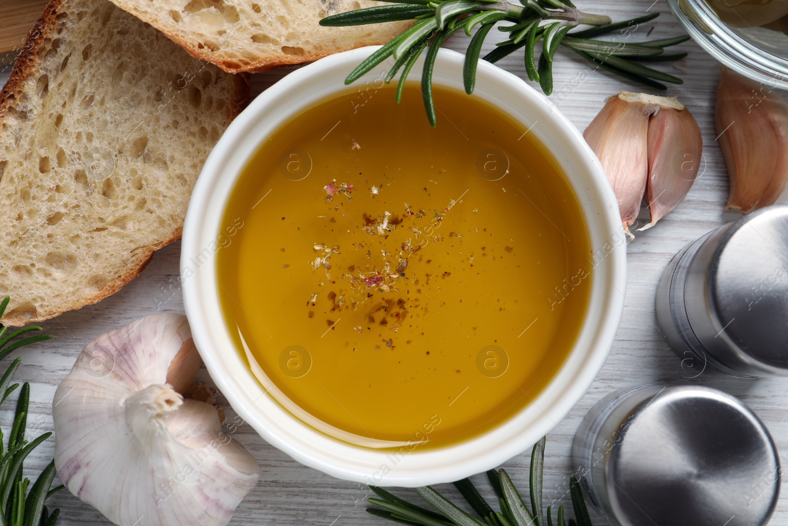 Photo of Bowl of fresh oil, bread, rosemary and garlic on white wooden table, flat lay