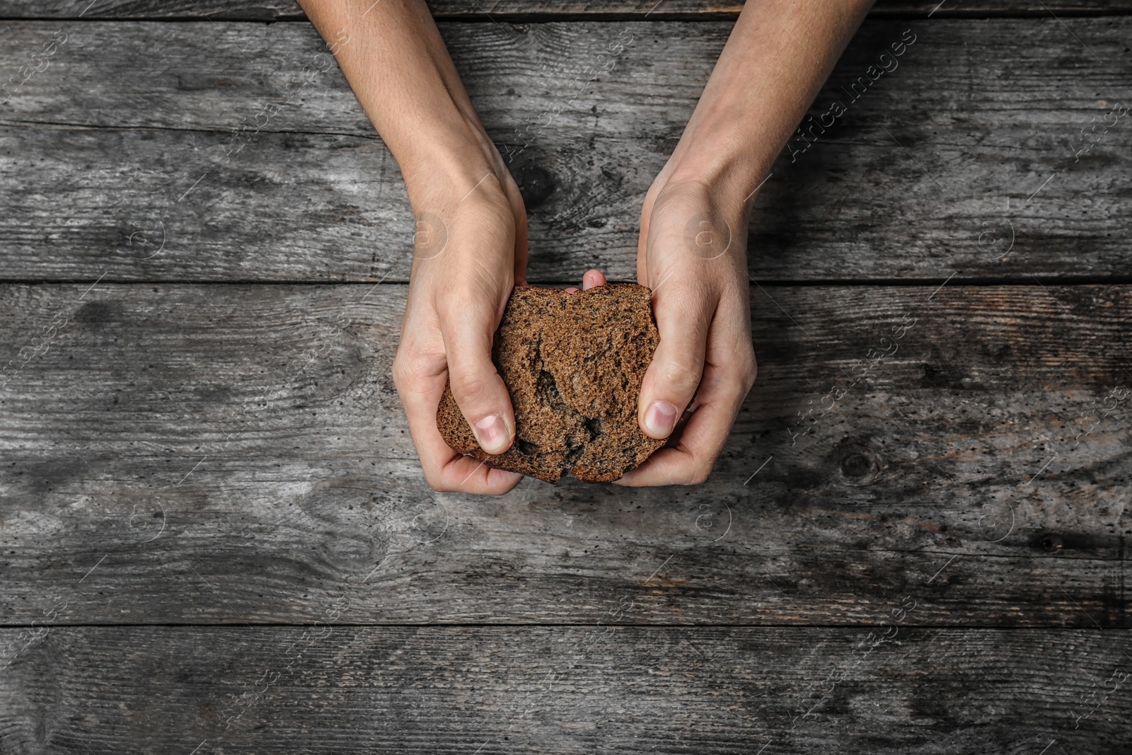 Photo of Poor woman holding piece of rye bread over wooden background, top view