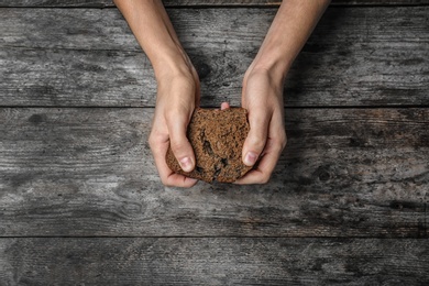 Poor woman holding piece of rye bread over wooden background, top view