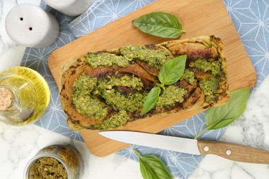 Freshly baked pesto bread with basil and knife on white marble table, flat lay