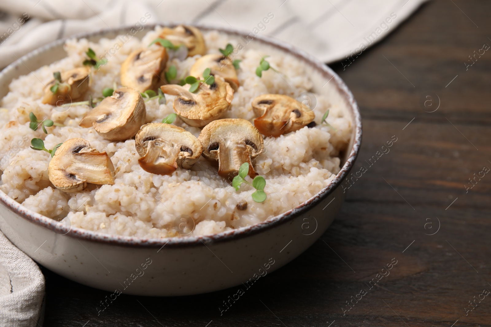 Photo of Delicious barley porridge with mushrooms and microgreens in bowl on wooden table, closeup