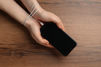 Photo of Woman holding smartphone in hands tied with charging cable at wooden table, top view. Internet addiction