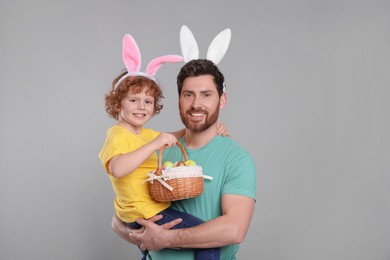 Photo of Happy father and son wearing cute bunny ears headbands on light grey background. Boy holding Easter basket with painted eggs