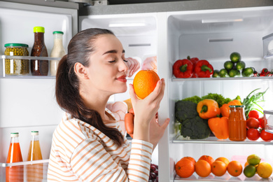 Photo of Young woman with orange near open refrigerator indoors