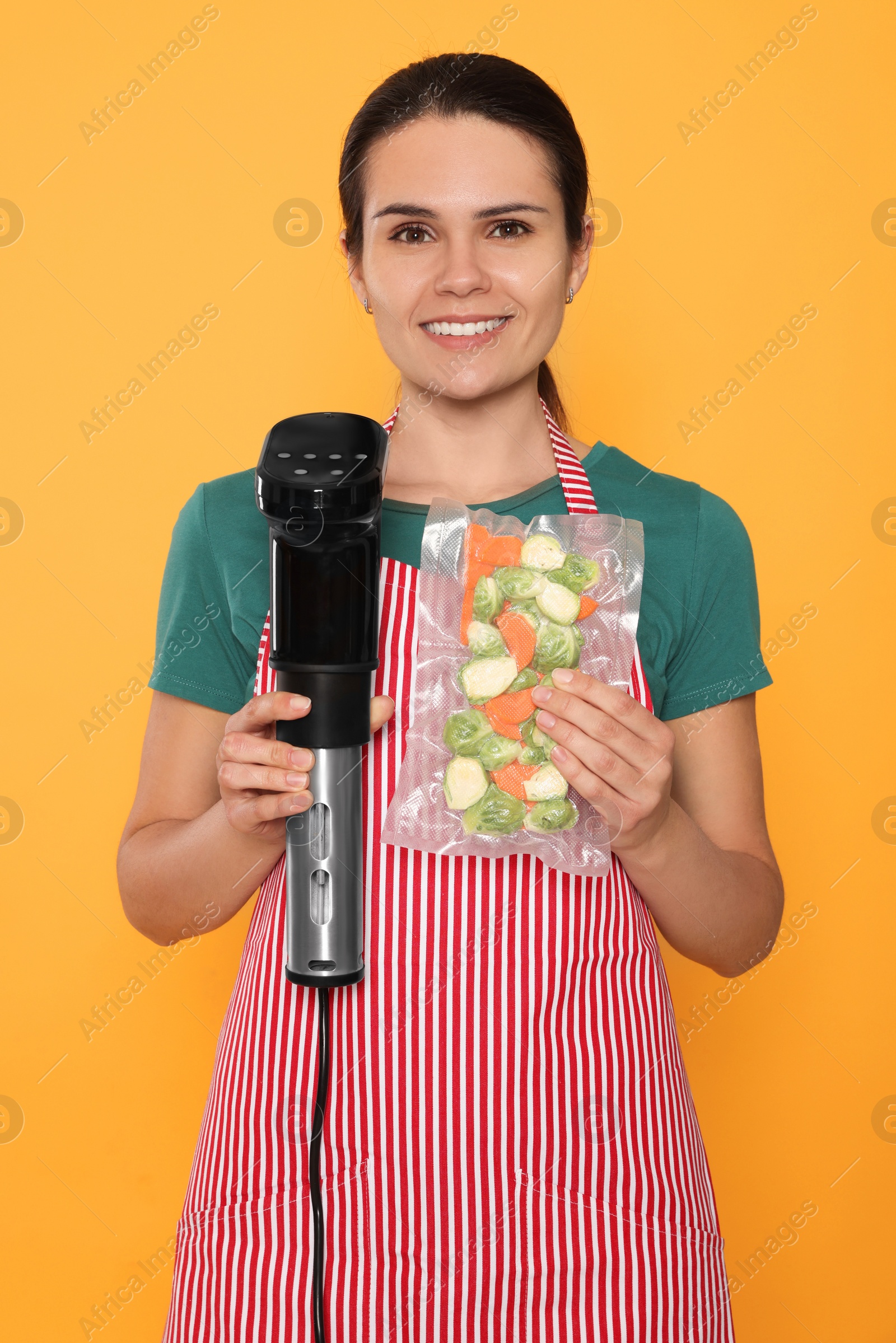 Photo of Beautiful young woman holding sous vide cooker and vegetables in vacuum pack on orange background