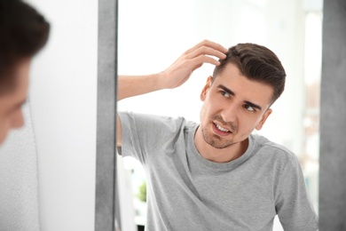 Young man with hair loss problem looking in mirror indoors