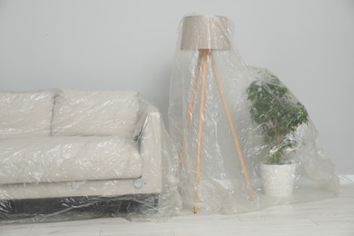 Sofa, lamp and houseplant covered with plastic film near light grey wall indoors