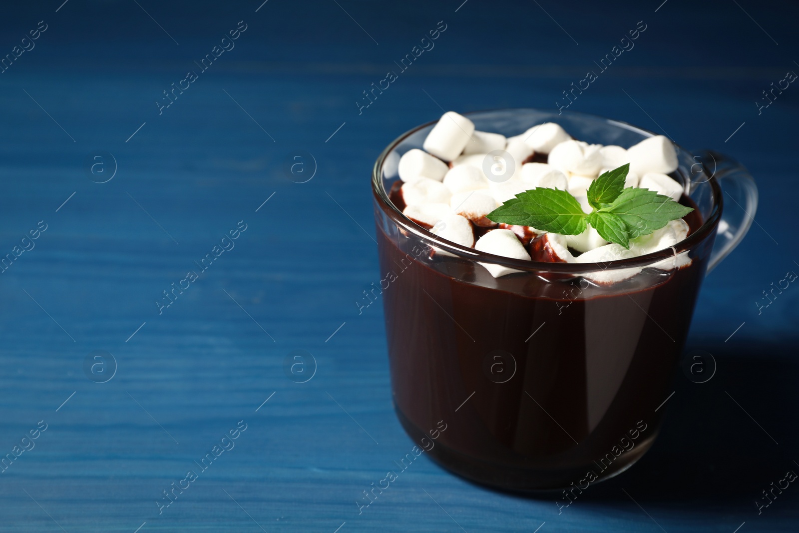 Photo of Glass cup of delicious hot chocolate with marshmallows and fresh mint on blue wooden table. Space for text