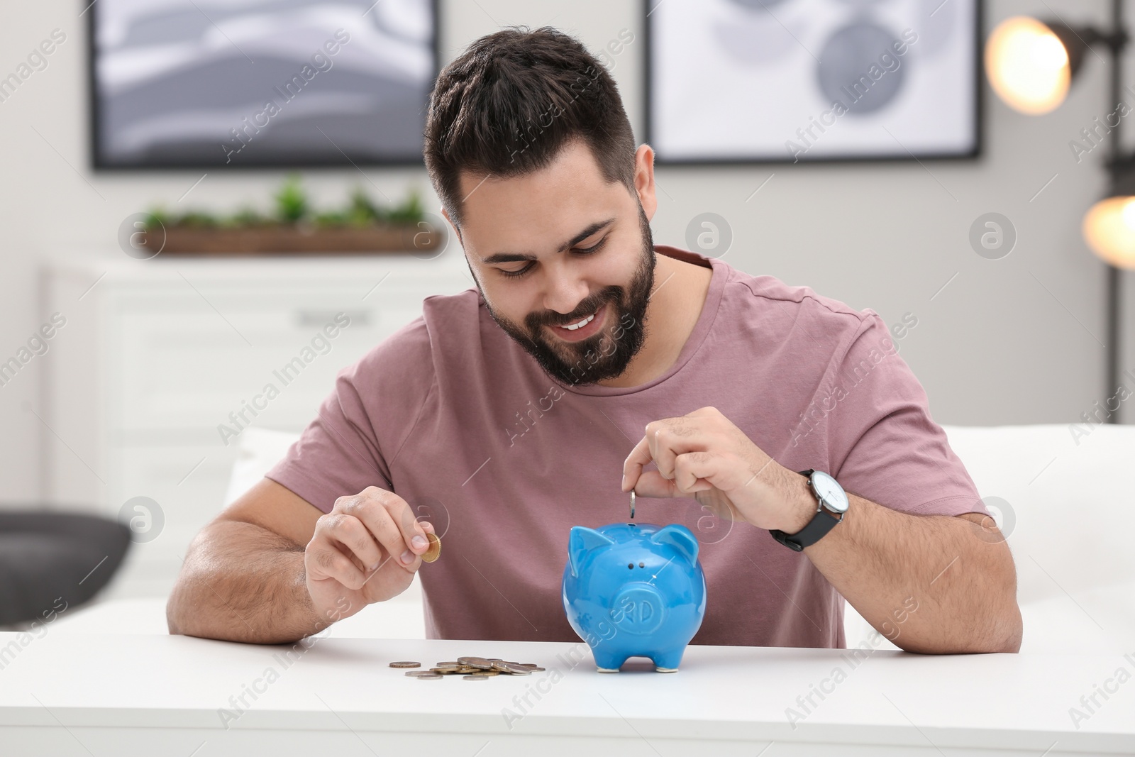 Photo of Happy young man putting money into piggy bank at white table indoors