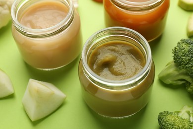 Jars with healthy baby food, broccoli and apple on light green background, closeup