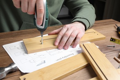 Photo of Man with electric screwdriver assembling wooden furniture at table, closeup