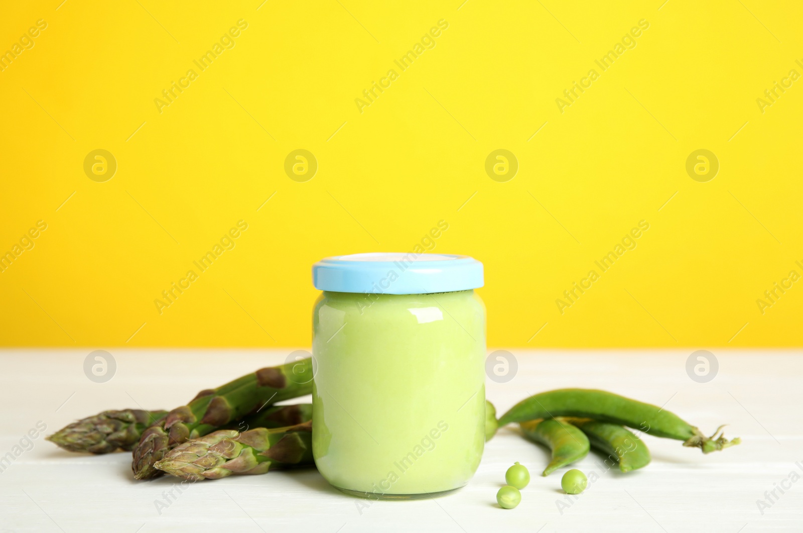 Photo of Jar with baby food, fresh pea pods and asparagus on white wooden table against yellow background