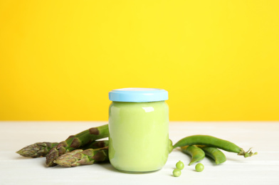 Photo of Jar with baby food, fresh pea pods and asparagus on white wooden table against yellow background