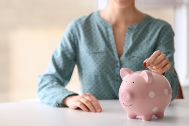 Photo of Woman putting coin into piggy bank at table indoors, closeup. Space for text