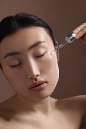 Beautiful young woman applying cosmetic serum onto her face on brown background