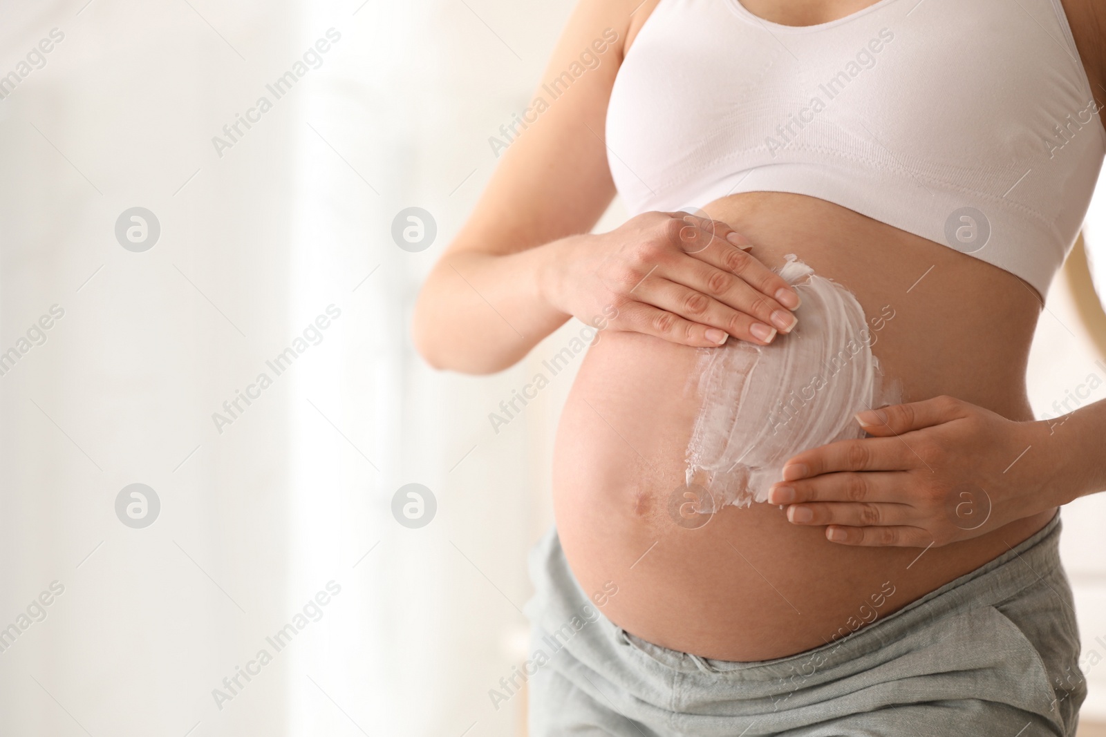 Photo of Young pregnant woman applying cosmetic product on belly in bathroom, closeup. Space for text