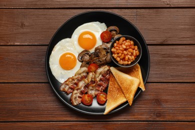 Plate with fried eggs, mushrooms, beans, bacon, tomatoes and toasted bread on wooden table, top view. Traditional English breakfast
