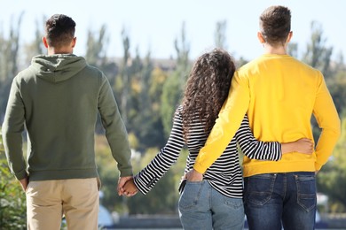 Photo of Woman holding hands with another man while hugging her boyfriend outdoors on sunny day, back view. Love triangle